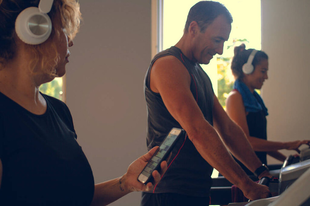 Drei Personen, die in einem Fitnessstudio Laufbänder benutzen und dabei Helme tragen, um Musik auf ihren Mobiltelefonen zu hören - Foto, Bild