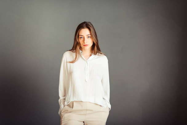 Νεαρή κοπέλα με λευκό πουκάμισο σε γκρι φόντο. Γαλλίδα με άσπρη μπλούζα πάνω σε γκρι τοίχους. Χωρίς ρετουσάρισμα. - Φωτογραφία, εικόνα