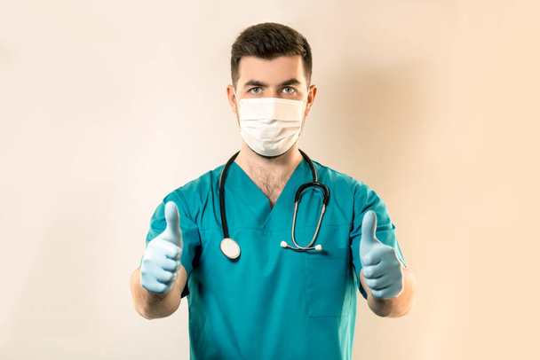 joven médico masculino en una máscara de virus muestra un pulgar hacia arriba con las manos
. - Foto, Imagen