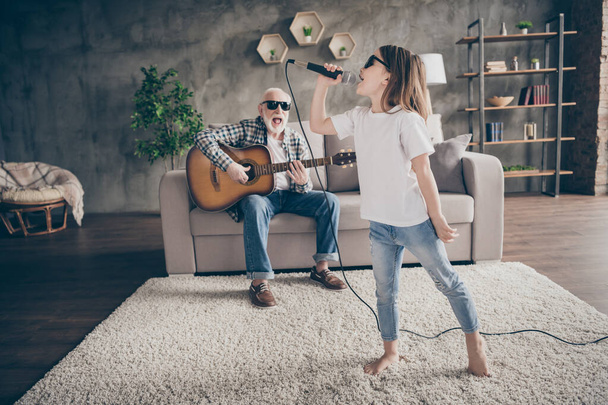 Photo of grandpa sit sofa play guitar little pretty granddaughter hold mic singing song spend time together cool trendy sun glasses stay home quarantine rejoicing modern living room indoors - 写真・画像