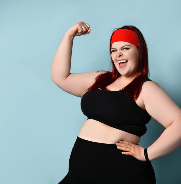 Fatty ginger lady in red headband, black top and leggings, fitness bracelet. Showing her muscles, smiling. Blue background - Foto, Bild