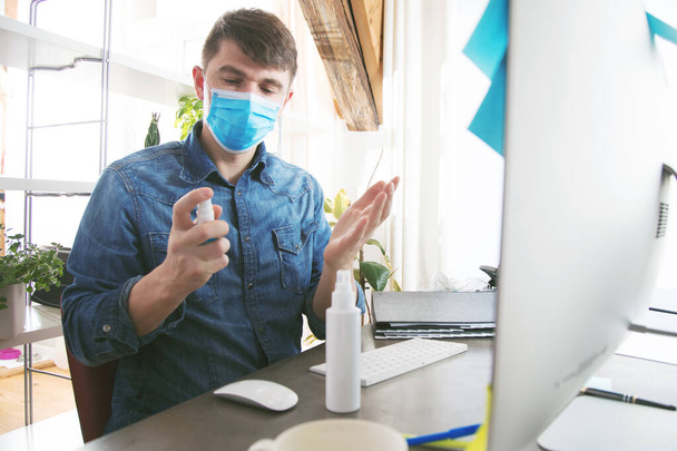 young man in medical mask cleaning hands with sanitizer while working in office - Foto, afbeelding