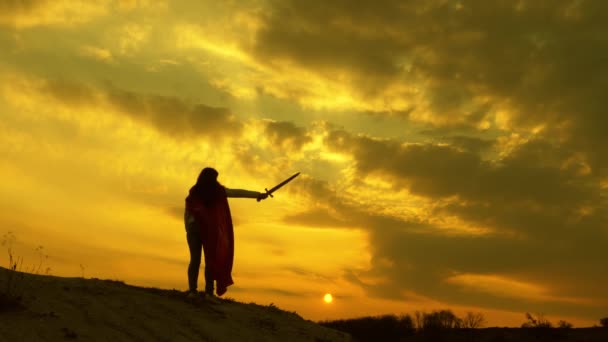 Elinde kılıçla ve kırmızı pelerinli süper kadın gün batımında bir dağın tepesinde duruyor. Özgür kadın süper kahramanı oynuyor. Kız, Roma Lenin 'ini güneşin parlak ışınlarıyla gökyüzüne karşı oynuyor. - Video, Çekim