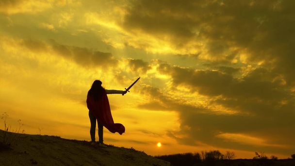 super kobieta z mieczem w ręku i w czerwonym płaszczu stoi na górze w świetle zachodu słońca. Wolna kobieta grająca superbohatera. dziewczyna gra Roman Lenin w jasnych promieni słońca przed niebem - Zdjęcie, obraz