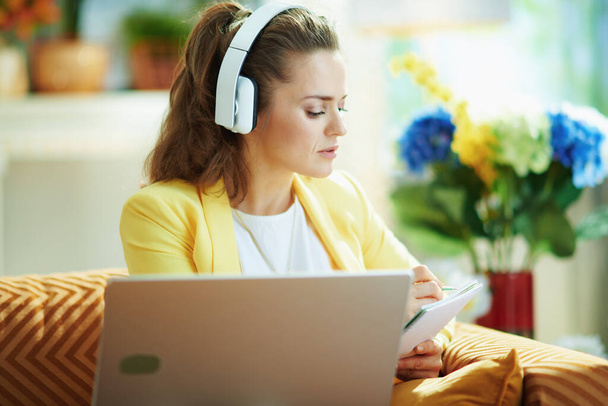 elegant middle age woman in jeans and yellow jacket with headphones subscribing to online education website in the modern house in sunny day. - Foto, Imagem