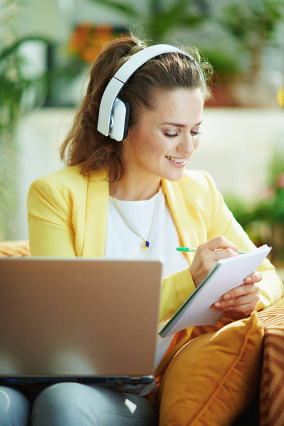 χαρούμενη κομψή γυναίκα με τζιν και κίτρινο σακάκι με ακουστικά βλέποντας μαθήματα internet στο σύγχρονο σπίτι σε ηλιόλουστη μέρα. - Φωτογραφία, εικόνα