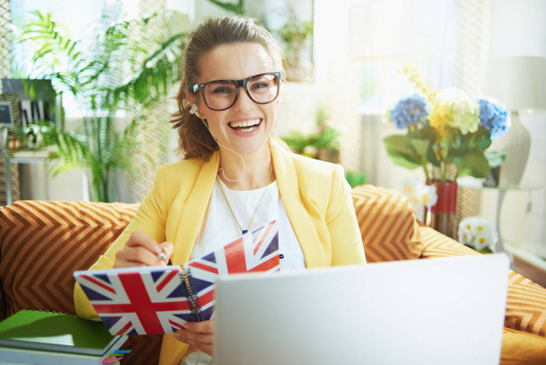 happy young woman in jeans and yellow jacket with laptop taking notes with a pen in a UK flag notebook at modern home in sunny day. - Fotoğraf, Görsel