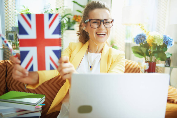 happy trendy learner woman in jeans and yellow jacket with laptop showing UK flag notebook at modern home in sunny day. - Photo, image