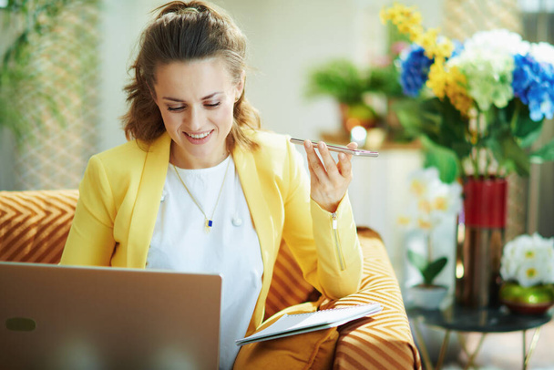 happy elegant 40 years old woman in jeans and yellow jacket with laptop and smartphone study online in the modern living room in sunny day. - Фото, изображение