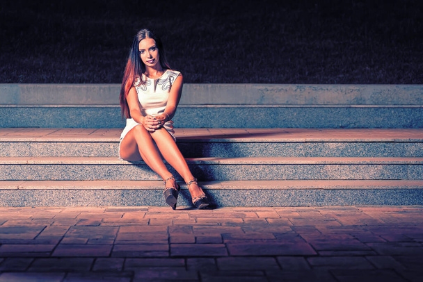 Woman in a short dress with long bare feet sat down to rest on the steps at night. She looks straight into the camera where her long legs are stretched. - Foto, immagini