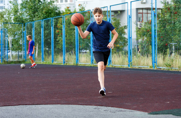 Aranyos mosolygós fiú kék pólóban kosarazik a városi játszótéren. Aktív tini élvezi szabadtéri játék narancs labdát. Hobbi, aktív életmód, sport gyerekeknek. - Fotó, kép