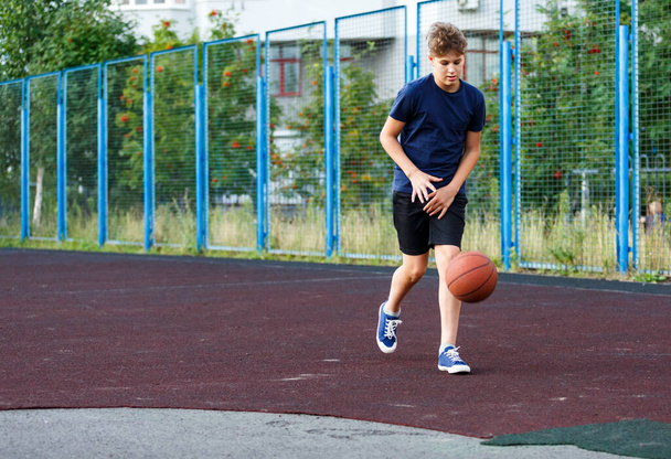Симпатичный мальчик в синей футболке играет в баскетбол на городской площадке. Активный подросток наслаждается игрой на свежем воздухе с оранжевым мячом. Хобби, активный образ жизни, спорт для детей, подростков. - Фото, изображение
