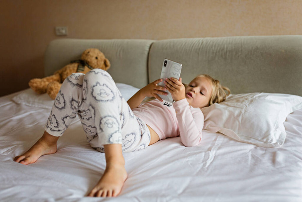 Cute little kid girl watching video on smartphone with smiley face alone on the bed, child using mobile phone with happy face at home. Stay at home quarantine coronavirus COVID-19 pandemic prevention - Fotografie, Obrázek