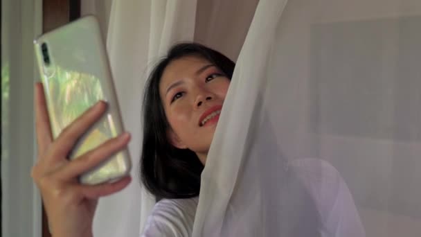 morning lifestyle shot on young beautiful and happy Asian woman in the morning having taking selfie with mobile phone at home terrace or hotel room balcony - Filmati, video