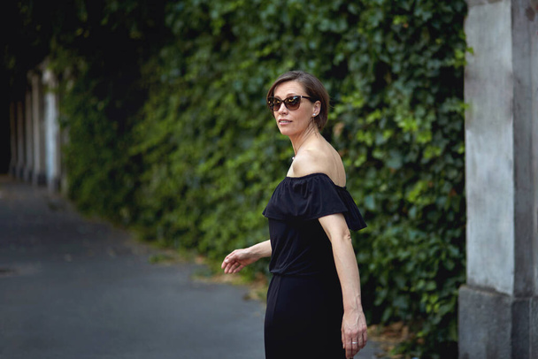 A woman in a black sexy dress with bare shoulders and sun-shy eyes walks along the wall of greenery. - Foto, immagini