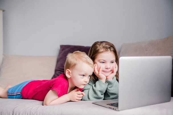 Twee kinderen een jongen en een meisje liggen op een bank in huiskleding en bekijken video 's op een laptop. Horizontale foto - Foto, afbeelding