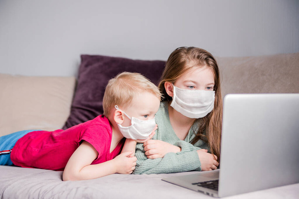 Zwei Kinder, Bruder und Schwester, liegen in medizinischen Masken auf dem Sofa, Laptop, häusliche Erziehung, bleiben zu Hause. Horizontales Foto - Foto, Bild