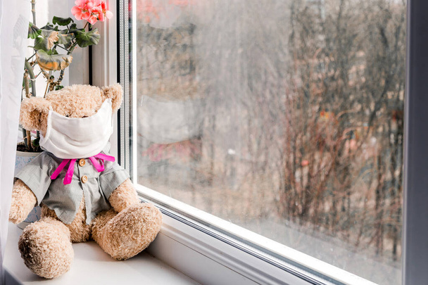 Niedźwiedź zabawka z białą maską twarzy patrząc przez okno z samoizolacji domowej kwarantanny podczas epidemii wirusa. Zdjęcie poziome - Zdjęcie, obraz