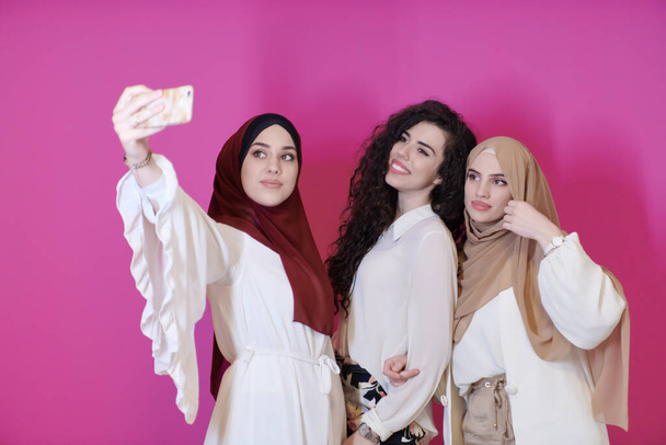 group of beautiful muslim women two of them in fashionable dress with hijab using mobile phone while taking selfie picture isolated on pink background representing modern islam fashion technology and ramadan kareem concept - Foto, Imagen