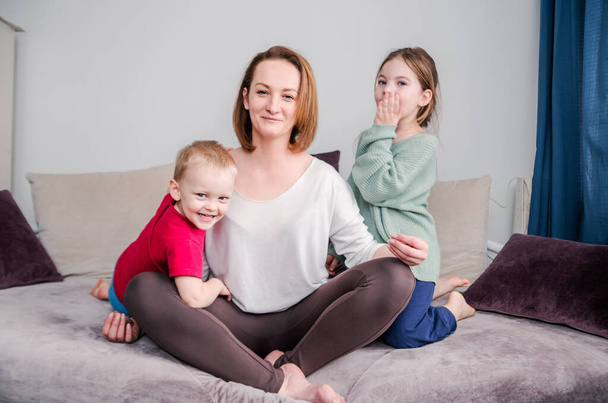 Η μαμά με τα δύο παιδιά της να κάθονται στον καναπέ στο σπίτι. Κοιτάζοντας την κάμερα και χαμογελώντας. Οριζόντια φωτογραφία - Φωτογραφία, εικόνα