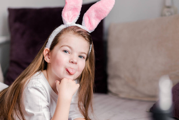 Little girl in a hoop in the form of rabbit ears lies on a sofa at home, posing for the camera and fooling around. Horizontal photo - Фото, изображение