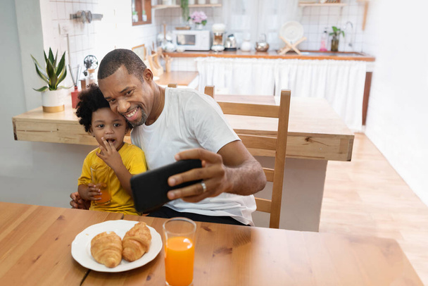 Happy African Father and son taking selfie photo with smartphone during lunch time in dining and kitchen room. Portrait smiling Dad and little kid boy making video call with mobile phone together in morning at home. Black Family having fun - Фото, изображение