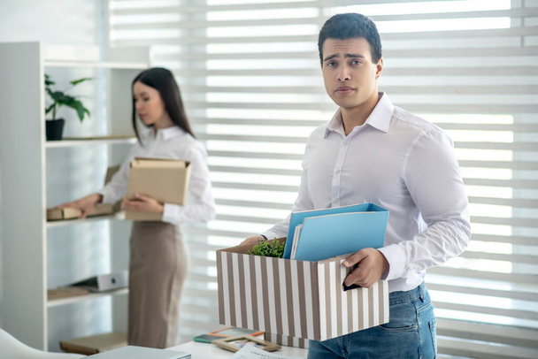 Upset young brunette male standing with the box of personal stuff, his female colleague standing behind him - Foto, Imagem