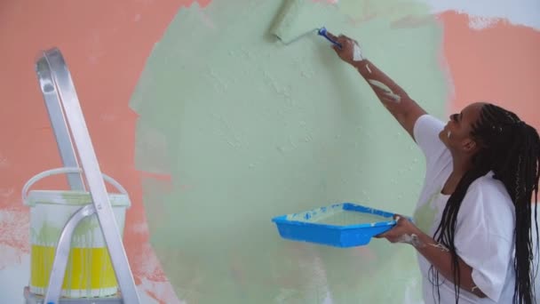 Koncepcja renowacji mieszkania. Uśmiechnięta Afroamerykanka przemeblowuje swój dom trzymając wałek malarski pokryty pomarańczową i zieloną farbą - Materiał filmowy, wideo