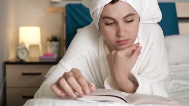 Girl is reading book. Attractive woman in bedroom in white bathrobe with towel on her head lies on stomach and reads book, turns over pages. Close-up - Záběry, video