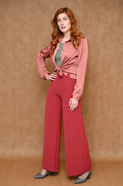 The model is dressed in a pink shirt and red pants, gray shoes. Full length vertical portrait straight Caucasian standing red hair pretty young woman on a beige background. - Foto, afbeelding