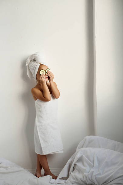 A young caucasian child with wrapped hair in white bath towels applying pieces of cucumber to her eyes on a white background - Foto, imagen