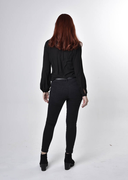 Portrait of a pretty girl with red hair wearing black jeans, boots and a blouse.  full length standing pose on a studio background, with back to the camera. - Fotografie, Obrázek