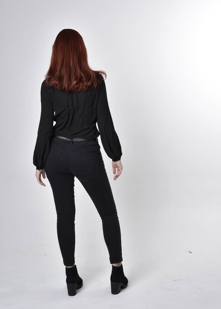 Retrato de una chica guapa de pelo rojo con vaqueros negros, botas y blusa. pose de pie de larga duración sobre un fondo de estudio, con la espalda a la cámara
. - Foto, imagen