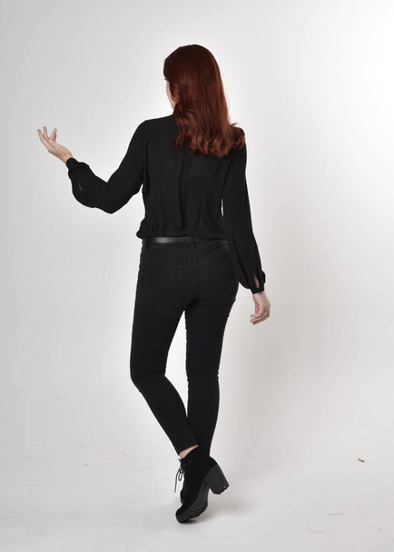 Retrato de uma menina bonita com cabelo ruivo vestindo calças de ganga pretas, botas e uma blusa. postura de pé de comprimento total em um fundo de estúdio, com volta para a câmera
. - Foto, Imagem