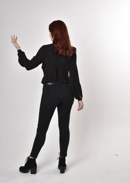 黒いジーンズ、ブーツ、ブラウスを着た赤い髪のきれいな女の子の肖像画。カメラに背を向けてスタジオの背景に立ち. - 写真・画像