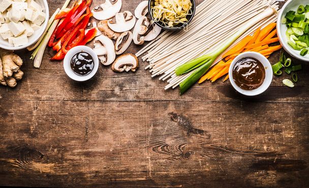 Asian vegetarian cooking ingredients for stir fry with tofu, noodles, ginger, cut vegetables, Sprout,green onion , lemongrass, hoisin and austern sauce on wooden rustic background, top view, border - Foto, afbeelding