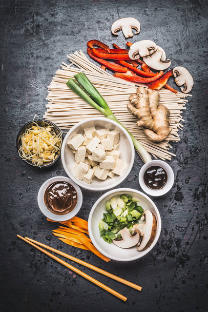 Verschiedene vegetarische asiatische Kochzutaten und Essstäbchen mit Tofu, Nudeln, Ingwer, geschnittenem Gemüse, Sprossen, grünen Zwiebeln, Hoisin und Austernsoße auf dunklem rustikalem Hintergrund, Draufsicht - Foto, Bild