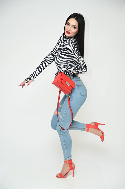 Model glamorous girl in blue jeans and a striped blouse stands right with a red bag on a white background. Vertical photo of a fashionable caucasian brunette. Side view. - Fotó, kép