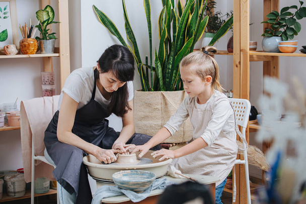 MIddle age brunette woman teaching little blond boy how to shape clay bowl on a pottery wheel in her private workshop. Chance to spent time on hobby during isolation. - Foto, Bild