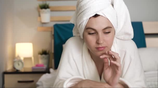 Girl is reading magazine. Attractive woman in bedroom in white bathrobe with towel on her head lies on stomach and turns over pages of fashion magazine. Close-up - Filmagem, Vídeo