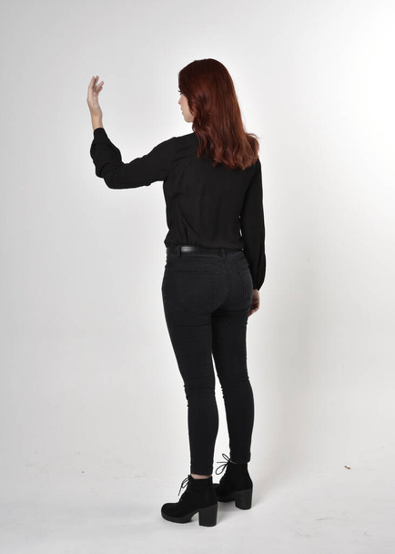 Portrét hezké dívky s rudými vlasy v černých džínách, botách a blůze. celková délka stojící póza na pozadí studia, se zády ke kameře. - Fotografie, Obrázek