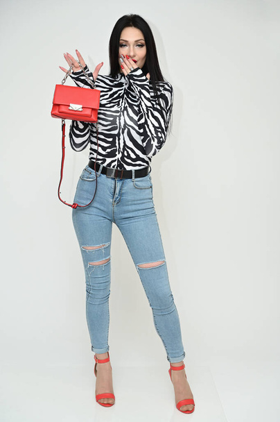 Full-length horizontal portrait of a fashionable caucasian brunette with excellent make-up. Glamorous girl in a striped blouse and blue jeans shows a red bag on a white background in the studio. - Photo, image