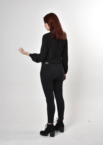 Portrait of a pretty girl with red hair wearing black jeans, boots and a blouse.  full length standing pose on a studio background, with back to the camera. - Foto, immagini
