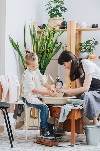 MIddle age brunette woman teaching little blond boy how to shape clay tableware on a pottery wheel in a private workshop. Chance to spend time on learning new skills during isolation. - 写真・画像