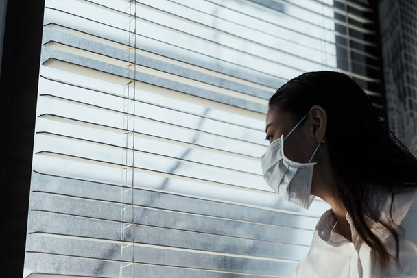 Siyah uzun saçlı kadın hijyenik bir maske takıyor camdan dışarı bakıyor.. - Fotoğraf, Görsel