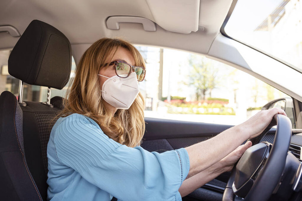 Shot of middle aged woman wearing face mask while driving her car during coronavirus pandemic.  - Valokuva, kuva