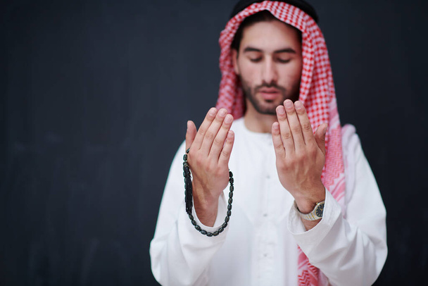 young arabian man in traditional clothes making traditional prayer to God, keeps hands in praying gesture in front of black chalkboard representing modern islam fashion and ramadan kareem concept - Photo, Image