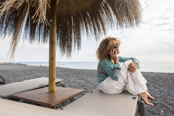 Feliz mulher caucasiana alegre fazer um telefonema sentado em uma espreguiçadeira na praia durante as férias de verão na moda na praia
 - Foto, Imagem
