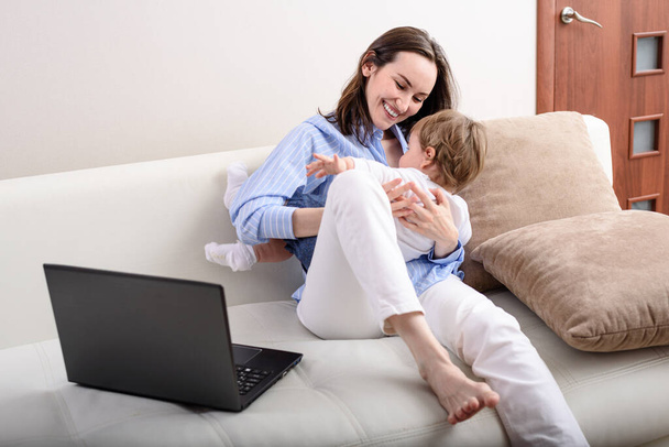 νεαρή ευτυχισμένη μαμά σε μπλε πουκάμισο παίζει με το παιδί στον καναπέ, άδεια μητρότητας, απομακρυσμένη εργασία στο φορητό υπολογιστή, έννοια της μητρότητας - Φωτογραφία, εικόνα