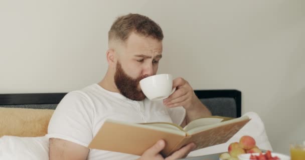 Hombre adulto leyendo historia y tomando café mientras está sentado con la bandeja llena de comida en sus piernas. Hombre barbudo guapo ordenando libro mientras desayuna en la cama. Concepto de ocio
. - Imágenes, Vídeo